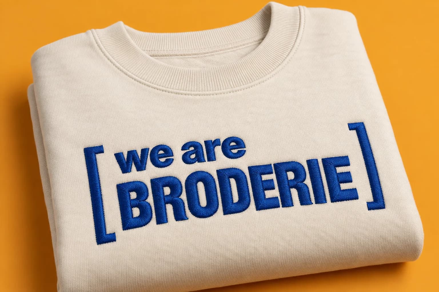 Sweat écru plié, brodé en point satiné bleu outremer du cadre « we are broderie », sur fond mandarine