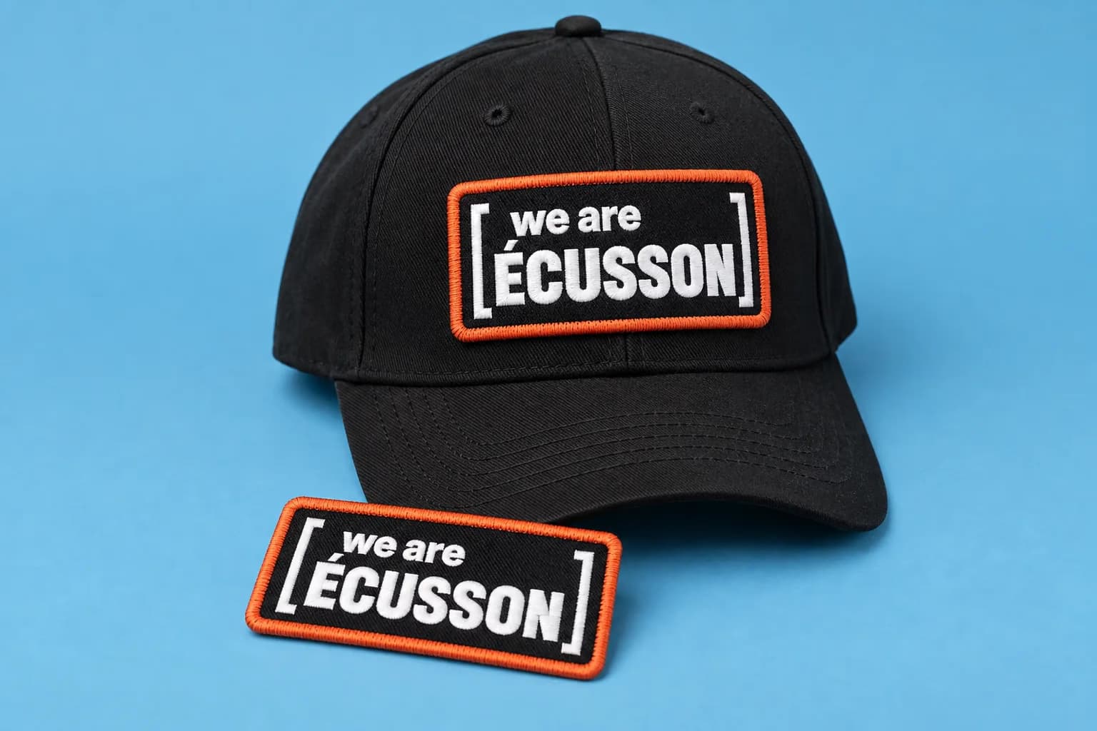 Casquette noire portant un écusson brodé bordé d'orange, un second écusson posé à côté, sur fond bleu ciel
